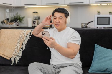 Evdeki Asyalı adam kablosuz kulaklık takıyor çalma listesinden hoşlanıyor ritme kafa sallıyor gözleri kapıyor ve gülümsüyor. Kanepede oturan Çinli adam yavaşça sallanıyor. Sakinliğini hisset, müzik çalarken dinleniyor.