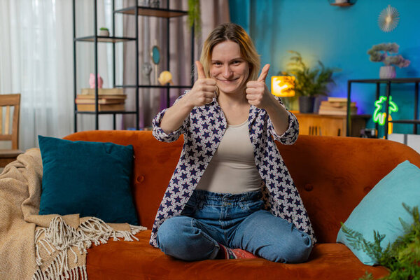 Portrait of excited smiling Caucasian woman looking approvingly at camera showing double thumbs up, like sign, good news, positive feedback. Happy blonde girl sitting on sofa in living room at home.