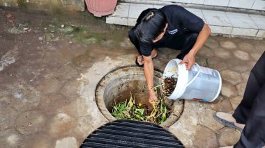Temizlik işçisi organik mutfak atıklarını açık hava gübre çukuruna atıyor. Sahne, Pekalongan Endonezya kentsel ortamında sürdürülebilir atık yönetimi ve çevre dostu uygulamaları yakalıyor, 13 Mayıs 2025