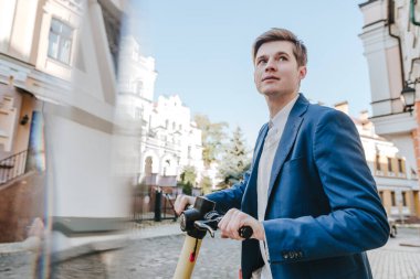 Şehir caddesinde elektrikli scooter kullanan takım elbiseli genç bir adam. Seçici odak.