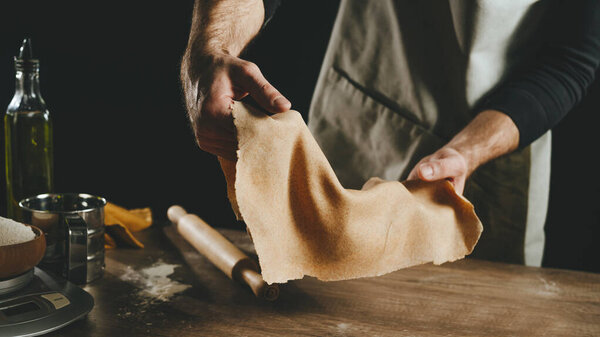 Unrecognizable man holding a thin dough against a dark background. Close-up of male hands preparing homemade noodles. Close-up of male hands preparing homemade noodles. Horizontal panoramic banner. Selective focus.