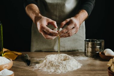 Karanlık duvara karşı ahşap arka planda hamur yoğuran tanınmaz bir adam. Yumurta kesen erkek ellerinin yakın çekimi. Ev yapımı eriştenin ikinci aşaması. Seçici odak.
