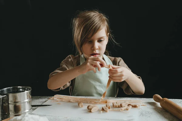 stock image Father and daughter in aprons holding dough cut into strips on a white table against a dark wall. Parent with child preparing homemade pastries or noodles. Selective focus.