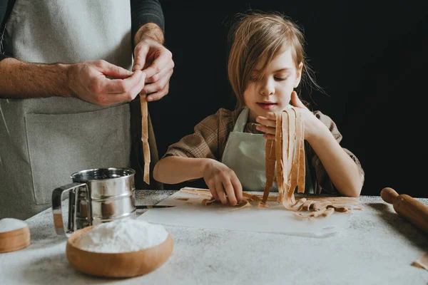 stock image Unrecognizable father and daughter in aprons holding dough cut into strips on a white table against a dark wall. Parent with child preparing homemade pastries or noodles. Selective focus.