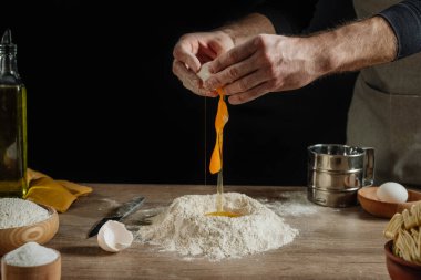 Karanlık duvara karşı ahşap arka planda hamur yoğuran tanınmaz bir adam. Erkek ellerinin yumurtaları kırdığı yakın plan. Ev yapımı eriştenin ikinci aşaması. Seçici odak.