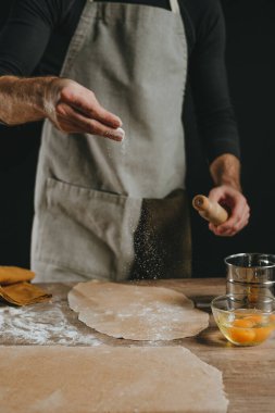 Tanınmayan bir adam ahşap arka planda merdaneyle hamura un serpiyor. Erkek eli yakın çekim, ev yapımı erişte hazırlıyor. Seçici odak.