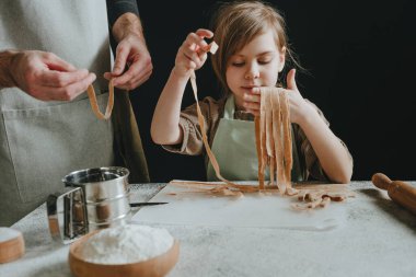 Önlüklü baba ve kızı, siyah bir duvara karşı beyaz bir masada kesilmiş hamur parçaları tutuyorlar. Çocuklu ebeveyn ev yapımı pasta ya da erişte hazırlıyor. Seçici odak.