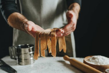 Önlüklü tanınmayan bir adam elinde hamurla karanlık bir duvara karşı beyaz bir masada şeritler halinde doğranmış. Erkek eli yakın çekim, ev yapımı erişte hazırlıyor. Seçici odak.