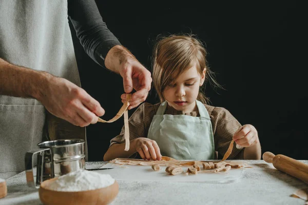 stock image Father and daughter in aprons holding dough cut into strips on a white table against a dark wall. Parent with child preparing homemade pastries or noodles. Selective focus.