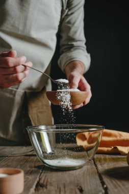 Önlüklü tanınmayan bir adam hamur pişirirken ahşap bir bardaktan cam bir kaba malzeme döküyor. Seçici odak.