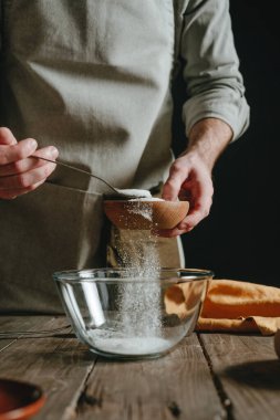 Önlüklü tanınmayan bir adam hamur pişirirken ahşap bir bardaktan cam bir kaba malzeme döküyor. Seçici odak.