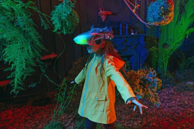 Ağaçların yanında duran sanal gerçeklik kulaklığı takan küçük bir kız. Doğal koşullarda arttırılmış gerçeklik oyunu. Gelecekteki teknoloji ve doğa arasındaki bağlantı kavramı. Seçici odak.