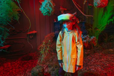 Parlak sanal gerçeklik kulaklığı takan küçük bir kız odada yosun, eğrelti otu ve kırmızı neon ışıklarda dallarla dikiliyor. Gelecekteki teknoloji ve doğa arasındaki bağlantı kavramı. Seçici odak.