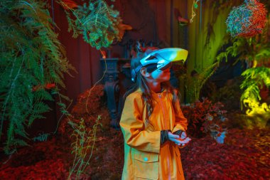 Parlak sanal gerçeklik kulaklığı takan küçük bir kız odada yosun, eğrelti otu ve kırmızı neon ışıklarda dallarla dikiliyor. Gelecekteki teknoloji ve doğa arasındaki bağlantı kavramı. Seçici odak.