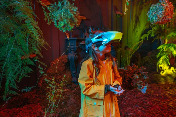 Parlak sanal gerçeklik kulaklığı takan küçük bir kız odada yosun, eğrelti otu ve kırmızı neon ışıklarda dallarla dikiliyor. Gelecekteki teknoloji ve doğa arasındaki bağlantı kavramı. Seçici odak.