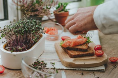 Erkeklerin elleri, ahşap zeminde balık ve domatesli sandviç yapan beyaz bir tencerede mikroyeşil turp kesiyor. Ev yapımı vitamin yiyeceği fikri.