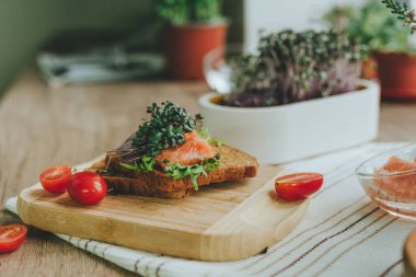 Tam buğday ekmekli sandviç, somon balığı, turp filizi mikroyeşil, ahşap zemin üzerinde domates. Seçici odak.