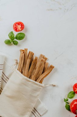 Domates ve fesleğen yanında pamuk torba içinde el yapımı çiğ makarna. Üst manzara, düz uzanma.