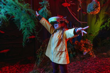 Little girl wearing luminous virtual reality headset standing near the trees. Augmented reality game in natural conditions. Concept of connection of the future technology and nature. Selective focus. clipart