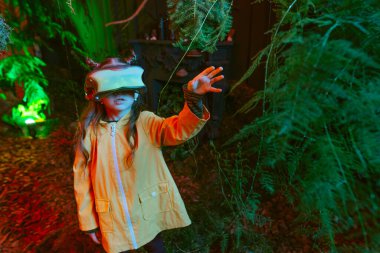 Parlak sanal gerçeklik kulaklığı takan küçük bir kız odada yosun, eğrelti otu ve kırmızı neon ışıklarda dallarla dikiliyor. Gelecekteki teknoloji ve doğa arasındaki bağlantı kavramı. Seçici odak.