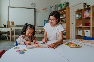 Kız ve öğretmen eğitici ahşap oyuncaklarla oynuyorlar ve anaokulunda geometrik şekilleri sınıflandırıyorlar. Seçici odak.