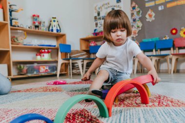 Anaokulunda yerde oturmuş gökkuşağı tahta oyuncağıyla oynayan küçük bir çocuk. Seçici odak.