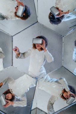 Beyaz elbiseli VR kulaklık takan, gri arka planda altıgen aynalarda poz veren genç bir kadın. Geometrik çerçevelerde gerçeküstü bir kadın portresi. Kaleydoskop etkisi.