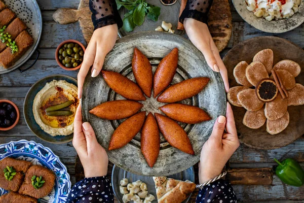 Hands of two women holding kibbeh plate . Turkish name is oruk or icli kofte . Dinner table with many meals on the table.
