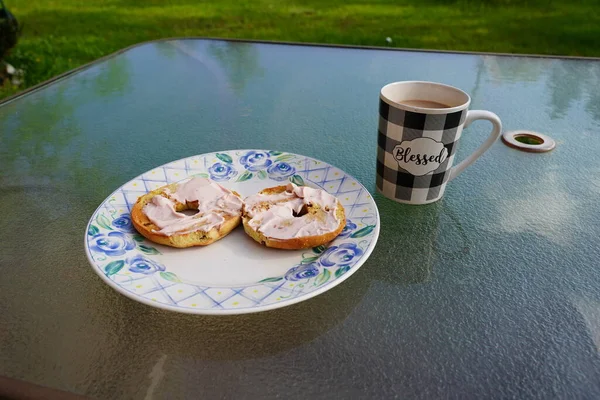 Cup of coffee and two bagels with cream cheese served outside on a glass table