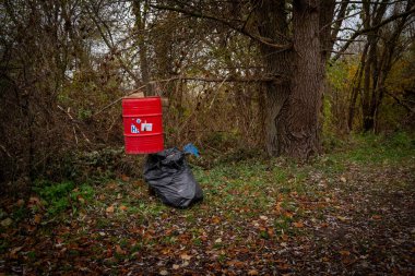 Otoparkta kırmızı bir çöp tenekesi var. Yanında da dolu bir çöp torbası.