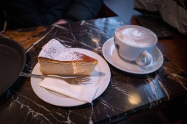 Masanın üzerinde bir parça cheesecake, bir bardak kakao ve bir fincan kapuçino var. 