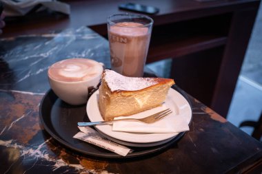 Masanın üzerinde bir parça cheesecake, bir bardak kakao ve bir fincan kapuçino var. 