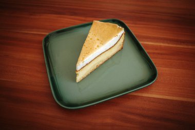 Altın kremalı leziz bir dilim peynirli kekin yakın çekimi, modern yeşil kare bir tabakta servis ediliyor, ahşap bir masanın üzerinde. Ev yapımı tatlı basit ve iştah açıcıdır. Fırın, kafe yaşam tarzı gibi temaları resmetmek için mükemmeldir.