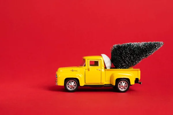 Yellow retro toy pickup carrying a Christmas tree on red background. Christmas and New Year celebration concept. Copy space, selective focus