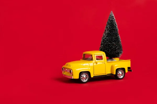 Yellow retro toy pickup carrying a Christmas tree on red background. Christmas and New Year celebration concept. Copy space, selective focus
