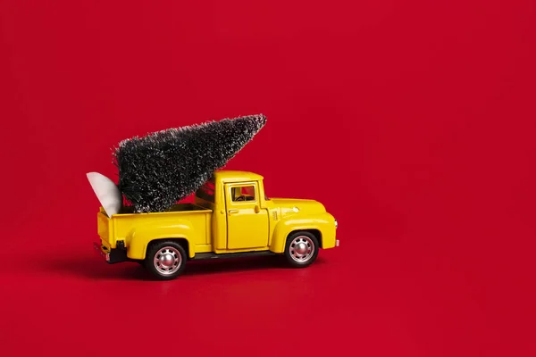 Yellow retro toy pickup carrying a Christmas tree on red background. Christmas and New Year celebration concept. Copy space,