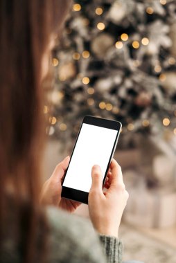 Mesaj atmak için yeri olan bir Noel modeli. Noel reklamı, uygulama şablonu. Işıklı güzel Noel ağacının arka planında boş ekranlı telefonu tutan eller..