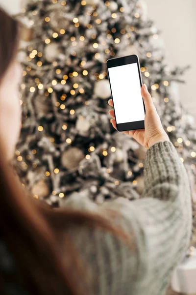 stock image Young woman congratulating and holding phone and looking at camera background of Christmas tree at cozy living room with festive interior. mock-up.