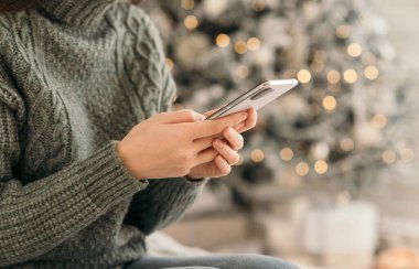 Genç bir kadın Noel kutlaması için cep telefonu kullanıyor. Çevrimiçi mesaj, bayram alışverişi, Noel kutlaması için akıllı telefon tutan kadınların yakın çekimleri. Yan görünüm
