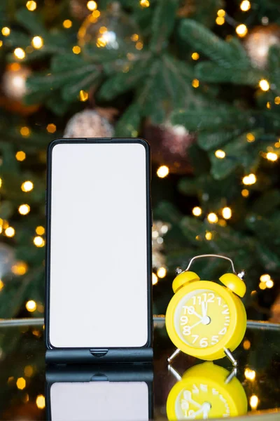 Phone with isolated screen and clock alarm on the background of the Christmas tree, bokeh and garland. New year gift, sales and offers
