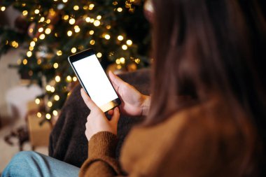Mesaj atmak için yeri olan bir Noel modeli. Noel reklamı, uygulama şablonu. Telefonun arkasında boş bir ekran olan el ele tutuşmuş altın gibi güzel bir Noel ağacının akşam ışıkları altında..