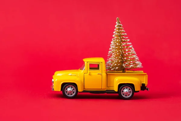 Yellow retro toy pickup carrying a Christmas tree on red background. Christmas and New Year celebration concept. Copy space