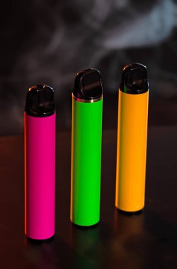 Siyah arka planda dumanlı farklı şekillerde tek kullanımlık renkli elektronik sigaralar. Modern sigara, buhar ve nikotin kavramı