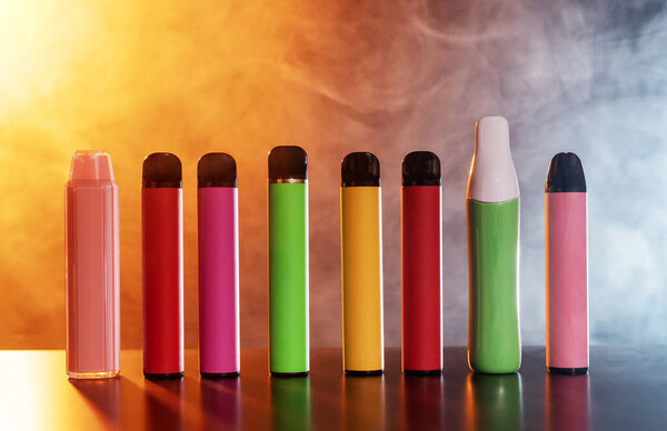 A set of colorful disposable electronic cigarettes of different shapes on a black background with smoke. Concept of modern smoking, vaping and nicotine