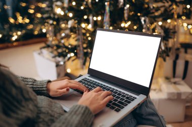 Kadın dizüstü bilgisayarda çalışıyor. Boş ekran ve taklit alanı var. Noel kutlaması için Noel ağacı ve çelenk ışıklarıyla dekore edilmiş ev.