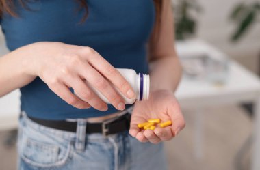 Genç kadın elinde sarı haplar, dişiyse şişeden takviye ve vitamin alıyor, yakın plan.