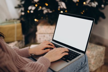 Kadın dizüstü bilgisayarda çalışıyor. Boş ekran ve taklit alanı var. Noel kutlaması için Noel ağacı ve çelenk ışıklarıyla dekore edilmiş ev. 