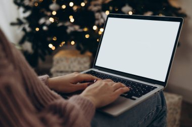 Kadın dizüstü bilgisayarda çalışıyor. Boş ekran ve taklit alanı var. Noel kutlaması için Noel ağacı ve çelenk ışıklarıyla dekore edilmiş ev. 