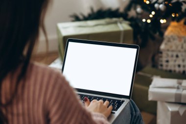 Kadın dizüstü bilgisayarda çalışıyor. Boş ekran ve taklit alanı var. Noel kutlaması için Noel ağacı ve çelenk ışıklarıyla dekore edilmiş ev. 