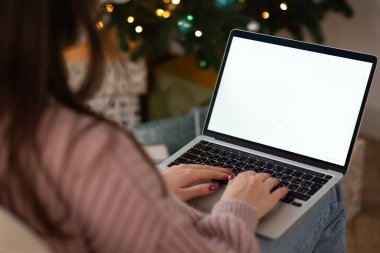 Kadın dizüstü bilgisayarda çalışıyor. Boş ekran ve taklit alanı var. Noel kutlaması için Noel ağacı ve çelenk ışıklarıyla dekore edilmiş ev. 